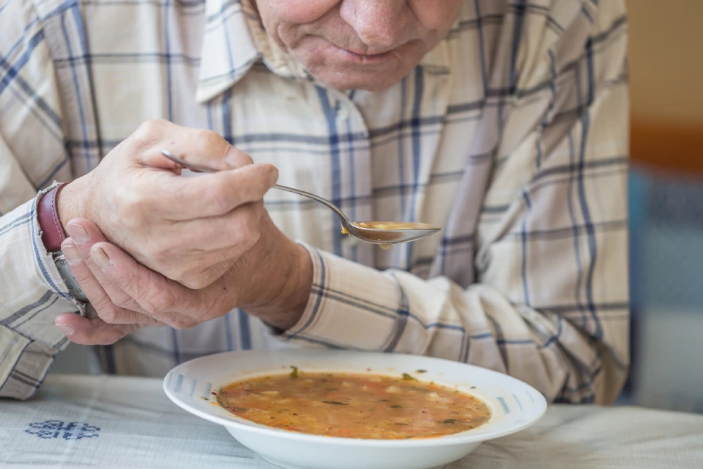 Man with Parkinson's disease eating soup while holding his wrist to keep is hand from shaking.