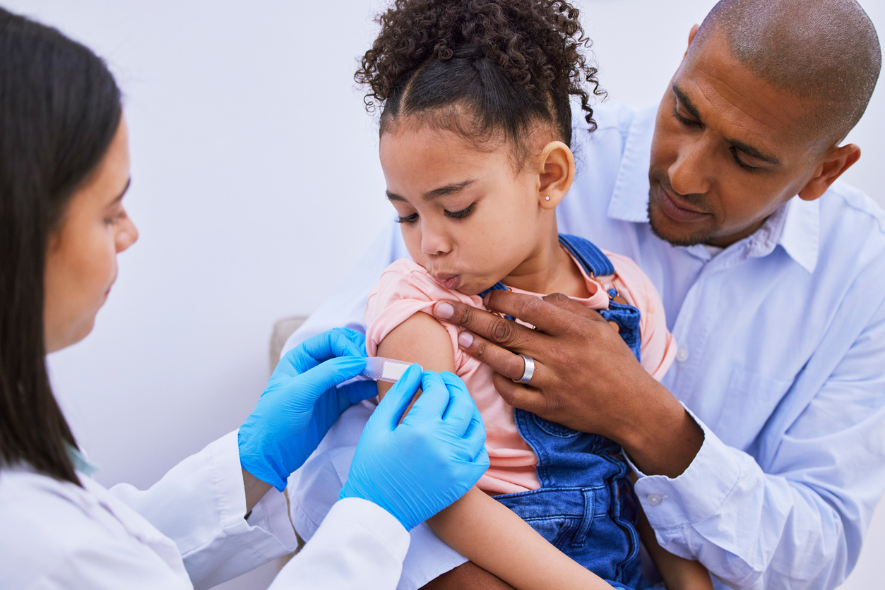 A father sits with his young daughter as a nurse places a bandage on her arm.