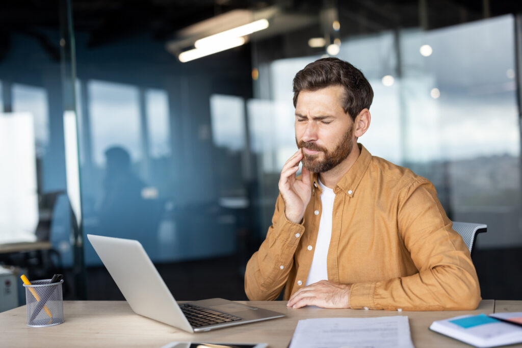 A young man sitting at his desk holds his jaw in pain.