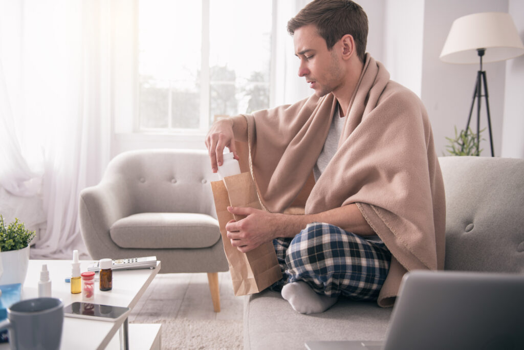 An ill young man is holding his prescription while he sits on the sofa covered with a blanket.