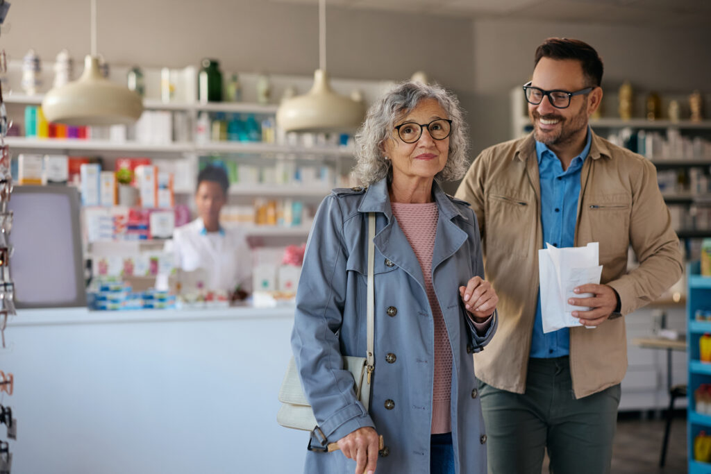 A son holds a prescription while escorting his elderly mother out of the pharmacy.