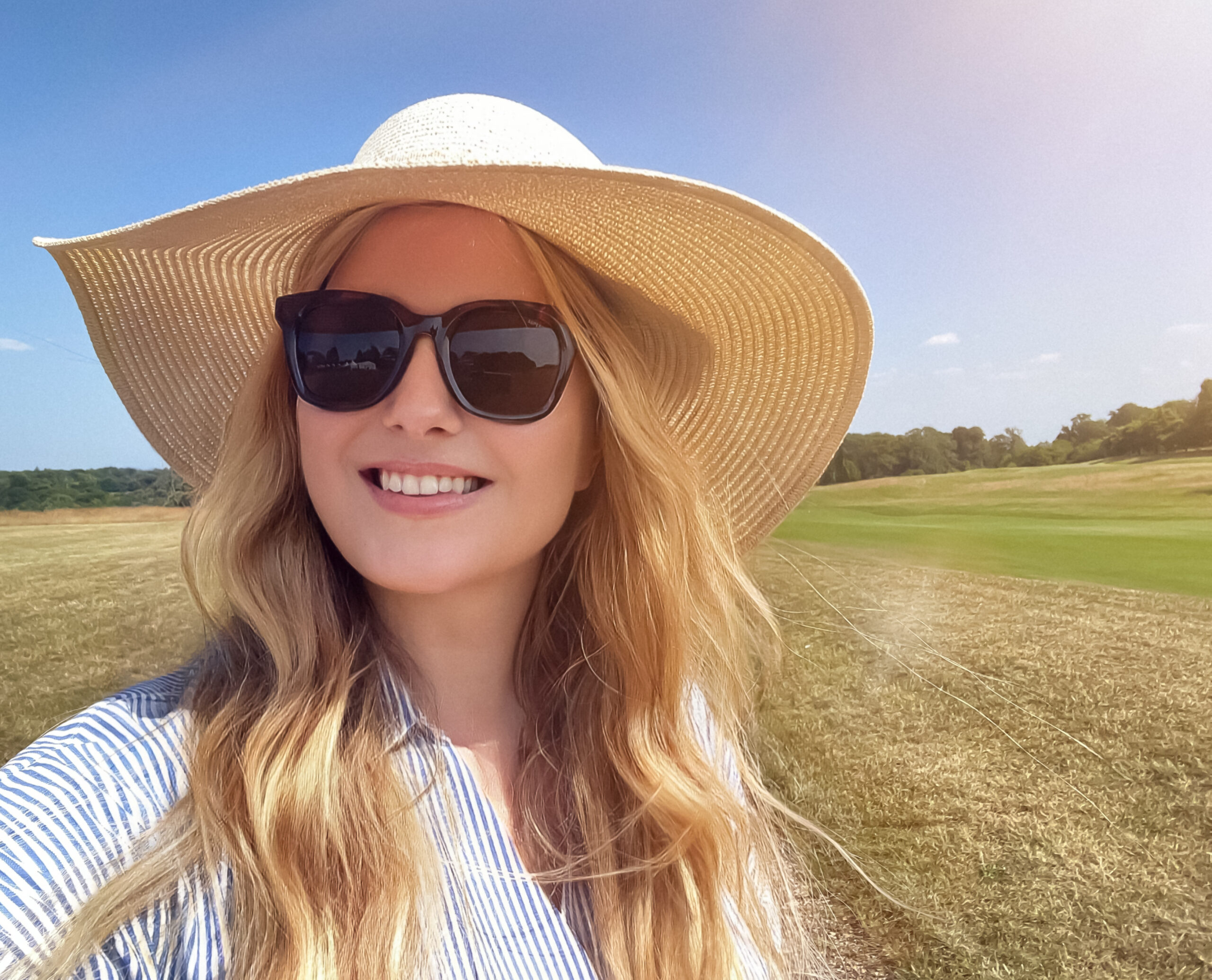 A woman wearing a wide-brimmed hat and sunglasses on a sunny day in a field.