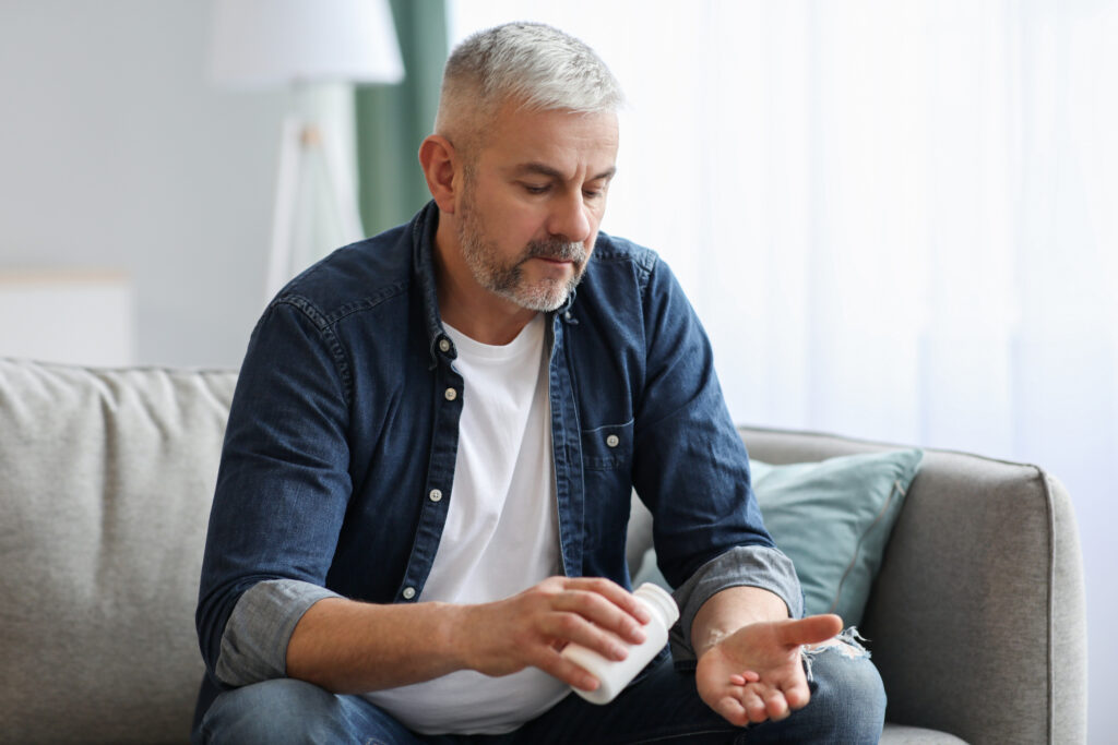 A man takes a couple tablets out of a pill bottle while he sits on the couch. 