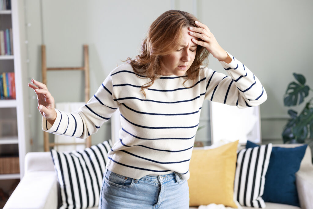 A woman is standing in her living room, holding her head in discomfort.
