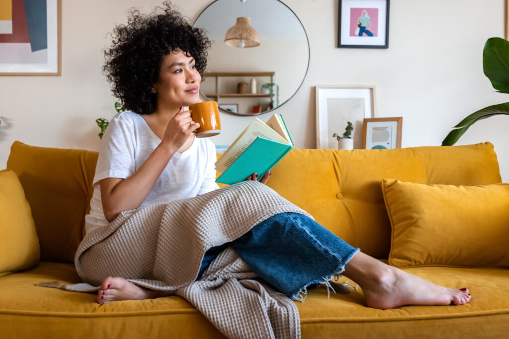 A young woman relaxes on the couch with a warm beverage and a book. 
