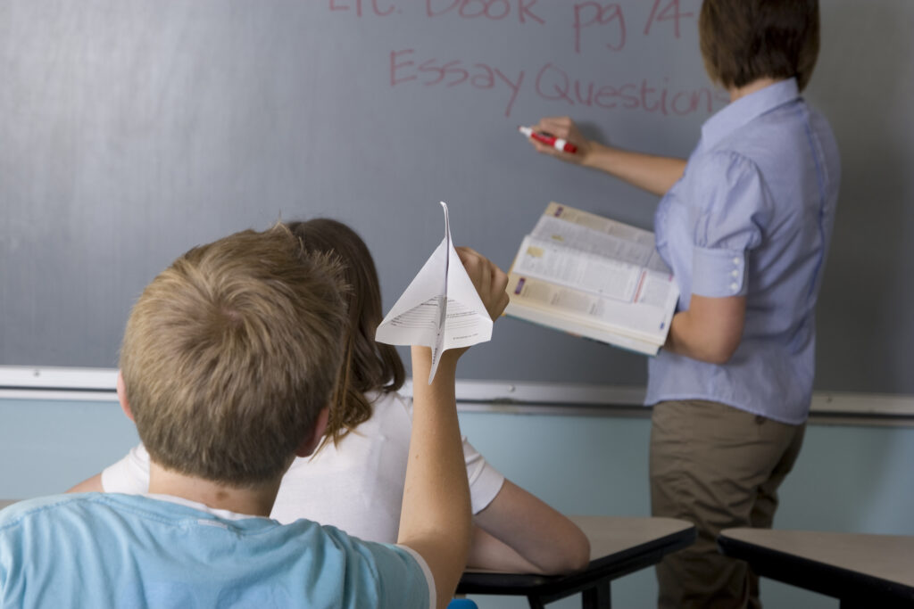 A young boy is about to throw a paper airplane at his teacher who's standing by the chalkboard. 